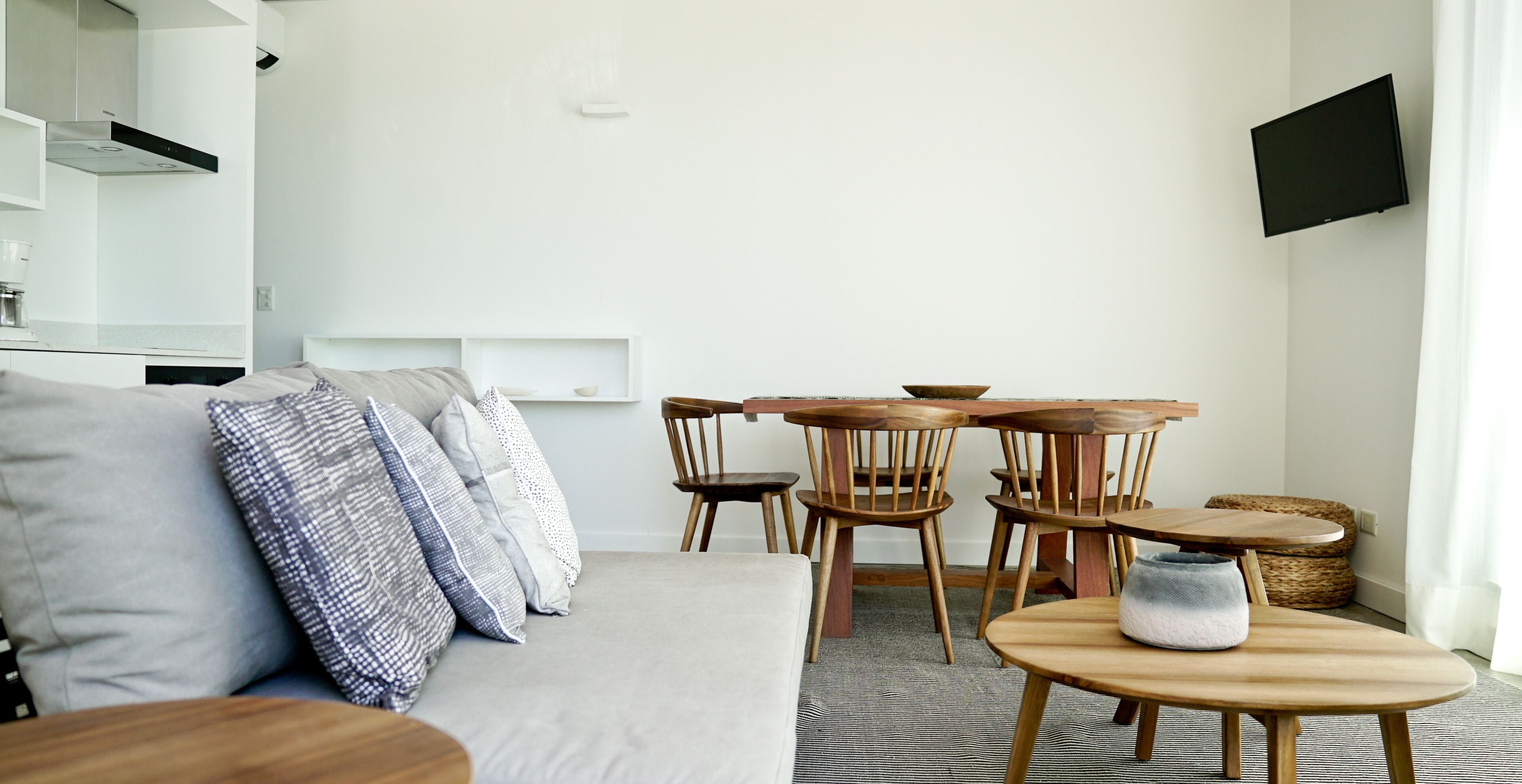 A modern living room with a grey sofa, wooden chairs, a round wooden table, and a flat-screen TV mounted on the wall.