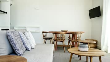 A modern living room with a grey sofa, wooden chairs, a round wooden table, and a flat-screen TV mounted on the wall.