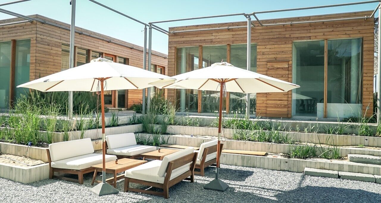 An outdoor seating area with wooden furniture and umbrellas, surrounded by planters and a modern building with large windows.