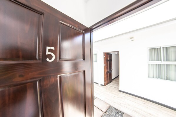 Une porte en bois portant le numéro 5, qui donne sur un couloir avec un mur blanc et une fenêtre ornée de rideaux.