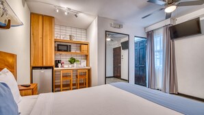 Habitación de hotel con una cama, armarios de madera, microondas, refrigerador, ventilador de techo y una ventana grande con cortinas.