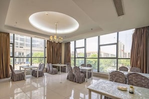 Um espaço de refeições de hotel com uma mesa central, várias cadeiras e grandes janelas com vista para a cidade.