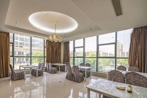 A spacious hotel dining area with large windows, a central chandelier, and multiple dining tables and chairs.