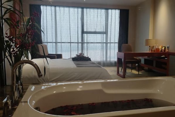 Habitación de hotel con una cama grande, un escritorio, una silla, una bañera con pétalos de rosa y un ventanal con cortinas traslúcidas.