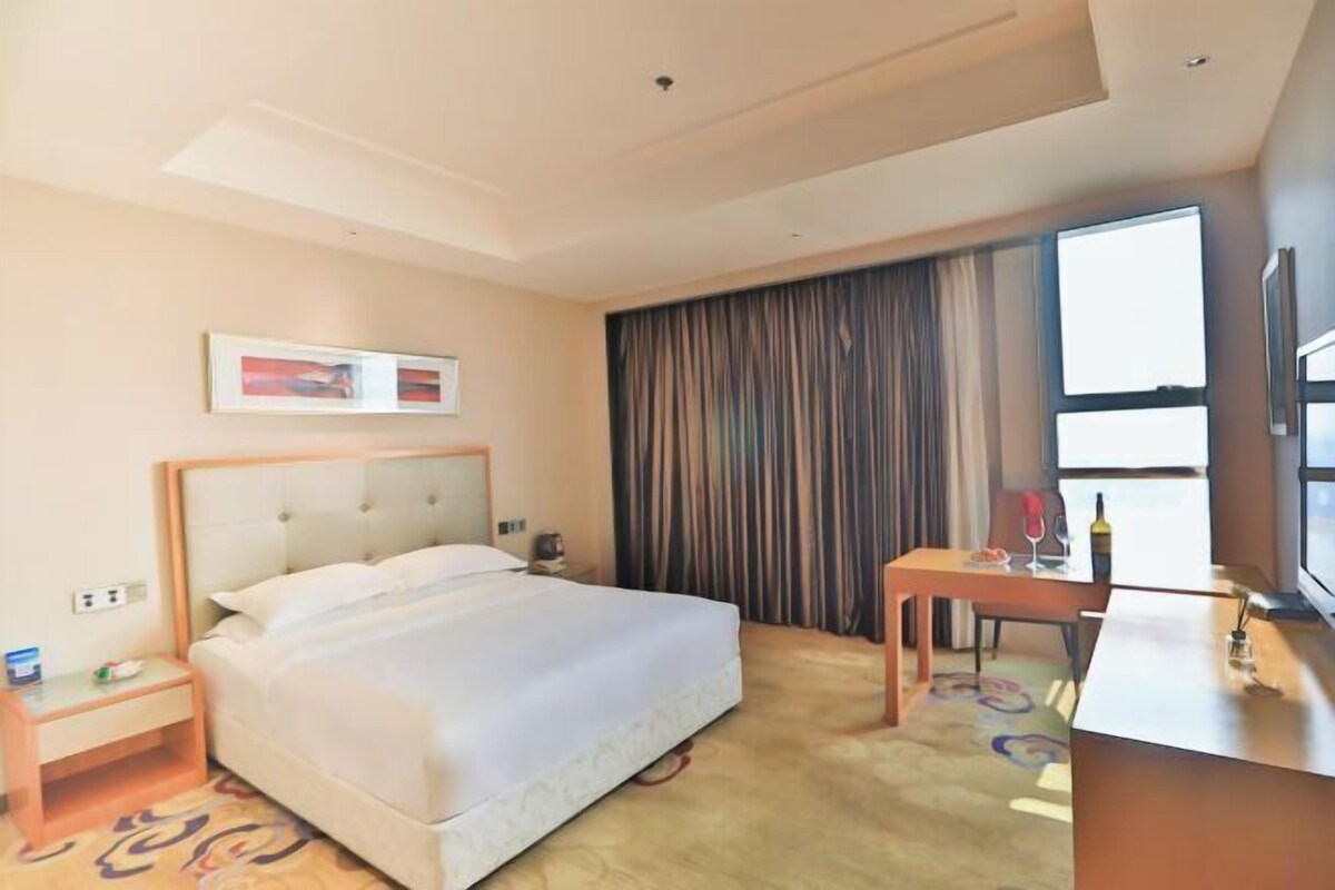 Una habitación de hotel con cama, escritorio, silla, televisión y ventana con cortinas.