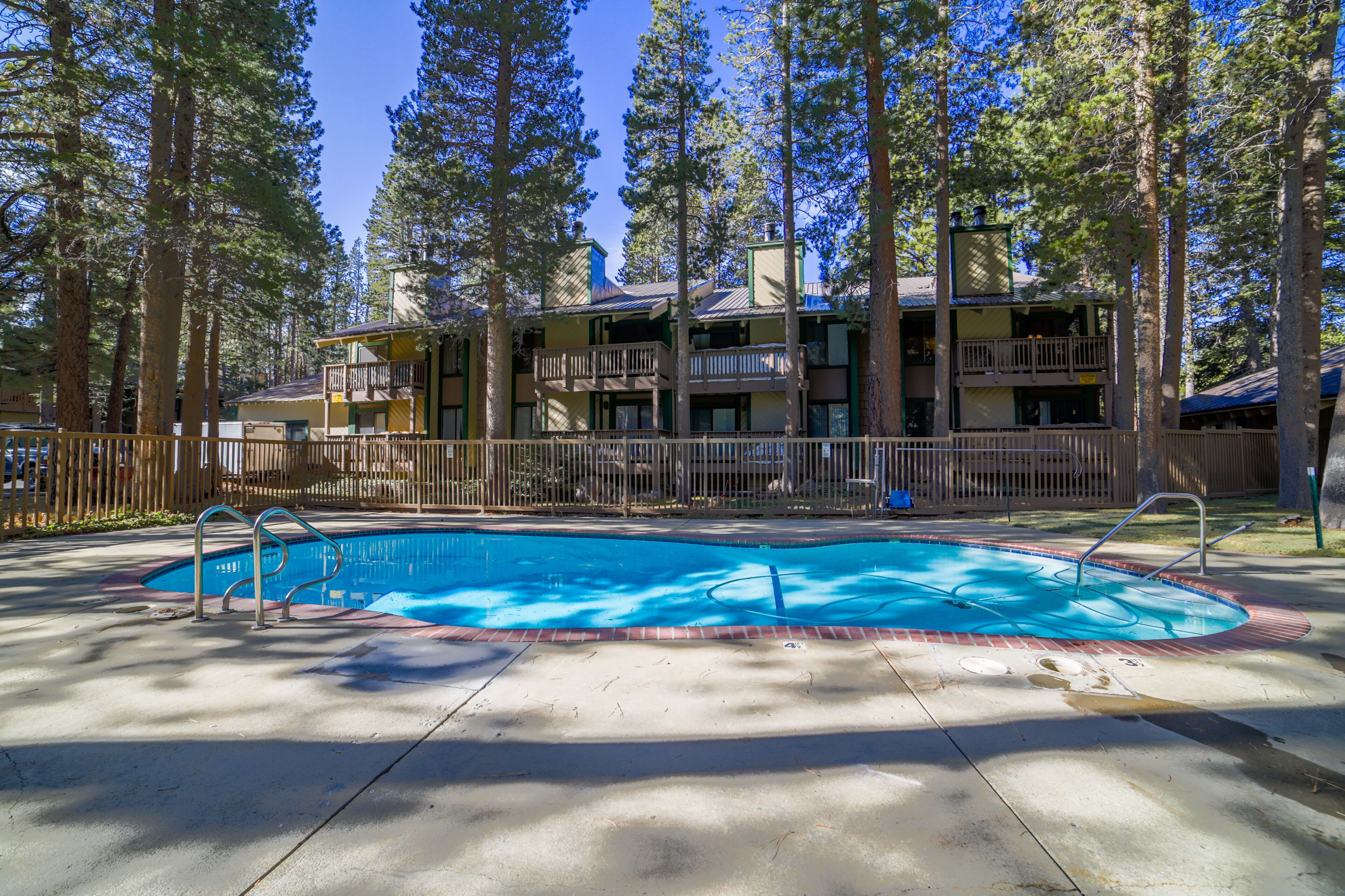 A swimming pool with a wooden deck, surrounded by tall trees and a multi-story building with balconies.