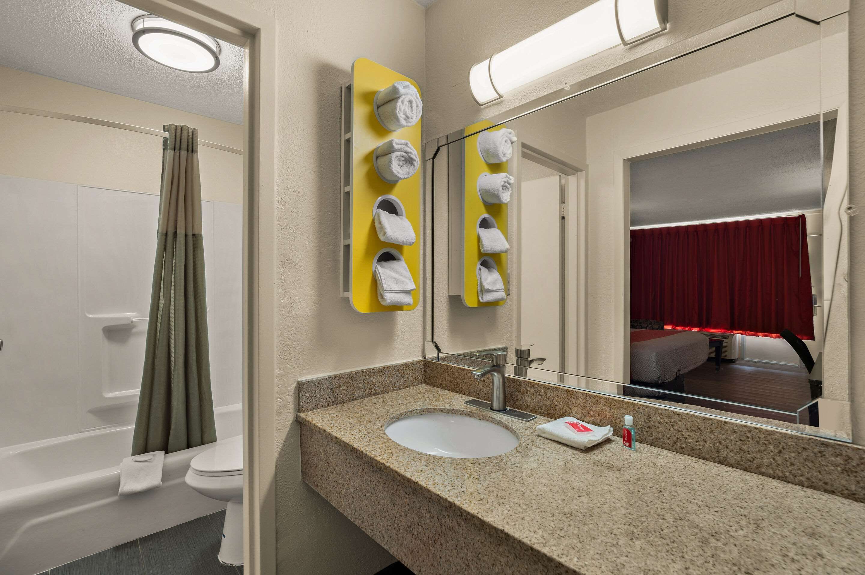 Un baño con lavamanos, espejo, dispensador de toallas y vista a un dormitorio con cortina roja.
