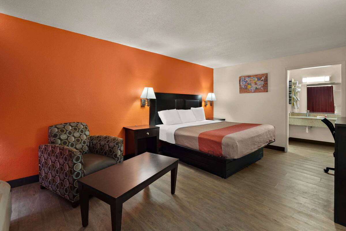 A hotel room with an orange accent wall, a bed with white bedding, two side tables with lamps, a small table, and a sofa.