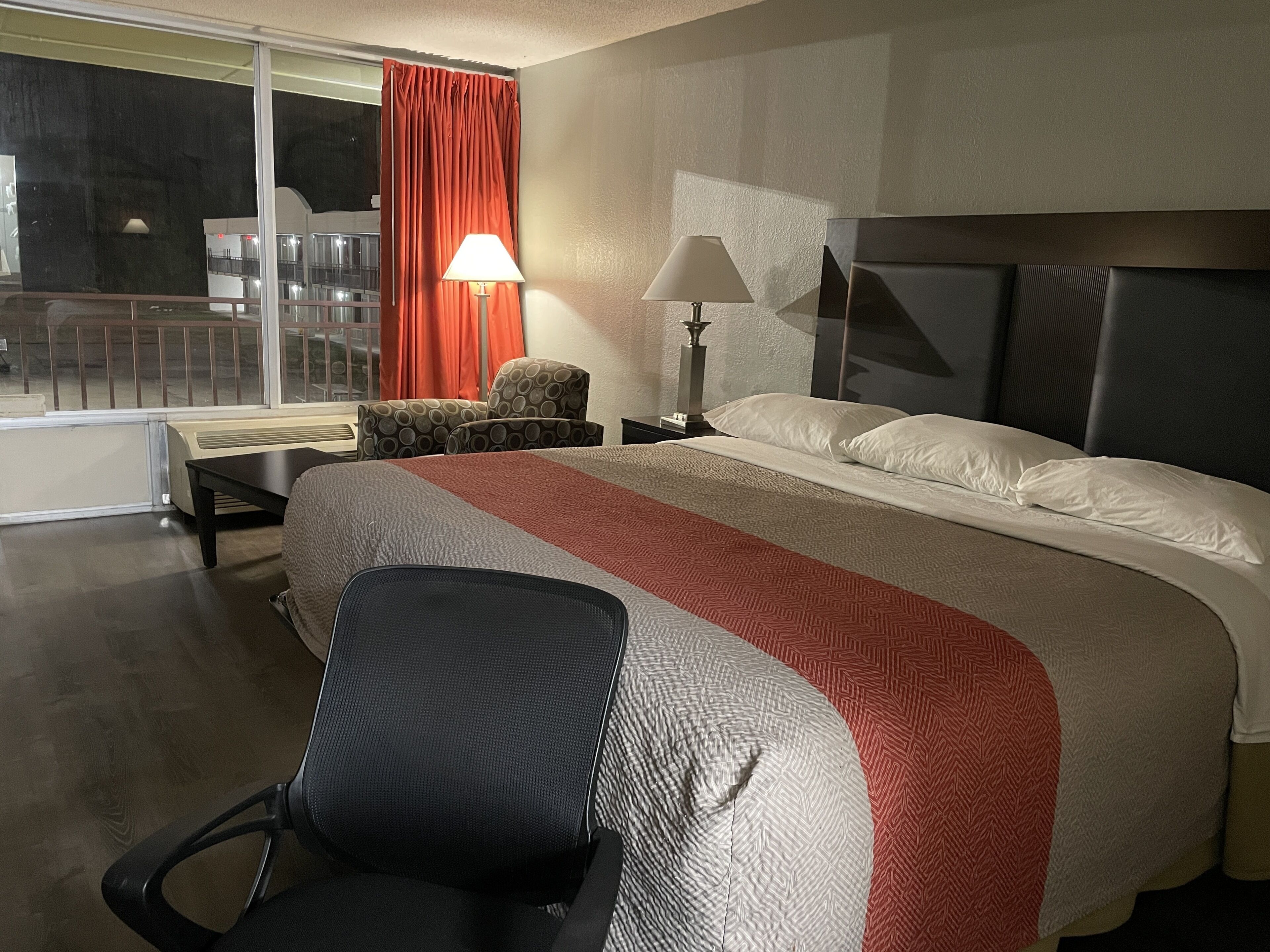 A hotel room with a large bed, a desk, a chair, a lamp, and a view of the outside.