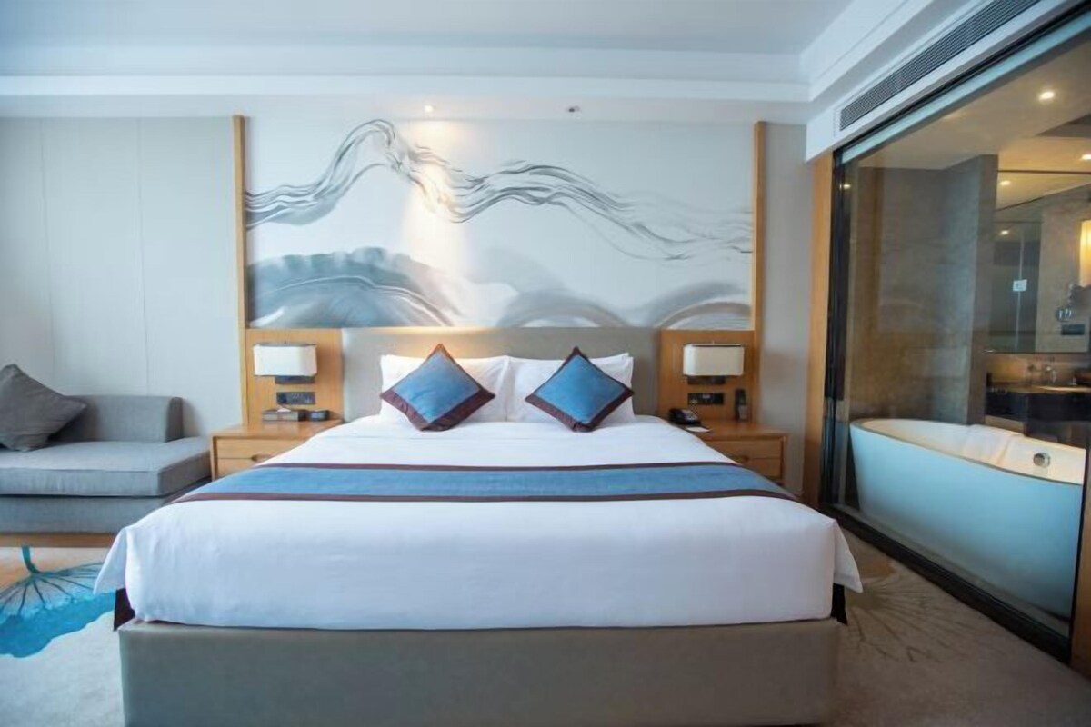 A hotel room with a large bed, two bedside tables with lamps, a sofa, and a large window with a view of a bathtub.