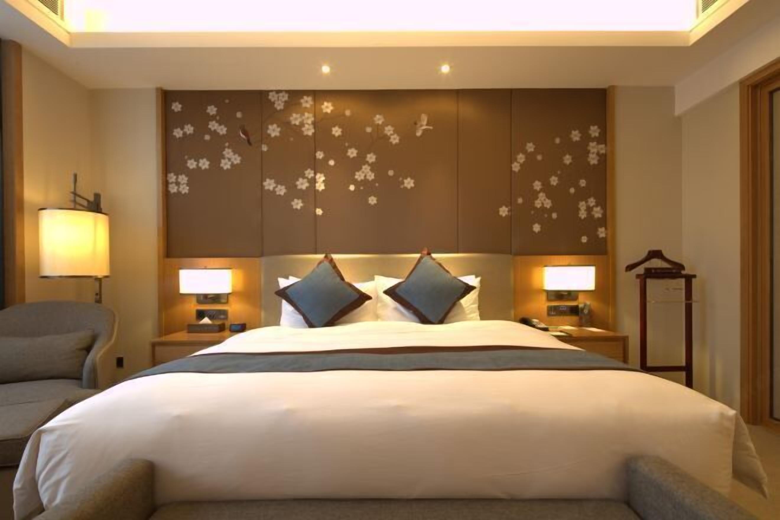 Habitación de hotel con una cama grande, dos lámparas de noche, una silla y un panel decorativo en la pared.