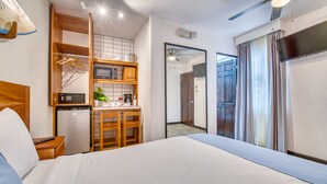 Ett hotellrum med en säng, ett litet pentry med mikrovågsugn, en kaffebryggare och ett litet kylskåp. Det finns en takfläkt och ett fönster med gardiner.