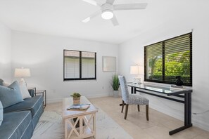 A modern living room with a sofa, a desk, and a chair. There is a ceiling fan, a window with blinds, and a plant.