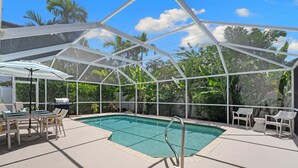 A covered outdoor swimming pool with lounge chairs and a table, surrounded by tropical vegetation.