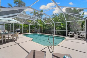 A covered outdoor swimming pool with lounge chairs and a table, surrounded by tropical vegetation.