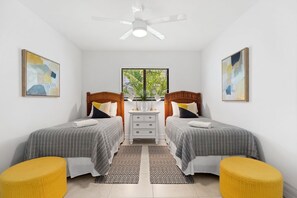 Quarto com duas camas de solteiro, cômoda, ventoinha no teto e janela com persianas.