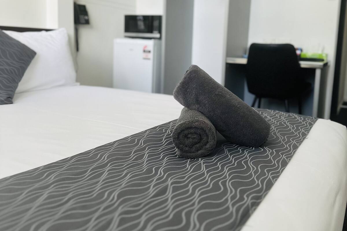 Cama bem arrumada com colcha estampada, toalha enrolada, travesseiro branco, escrivaninha pequena com cadeira e micro-ondas.