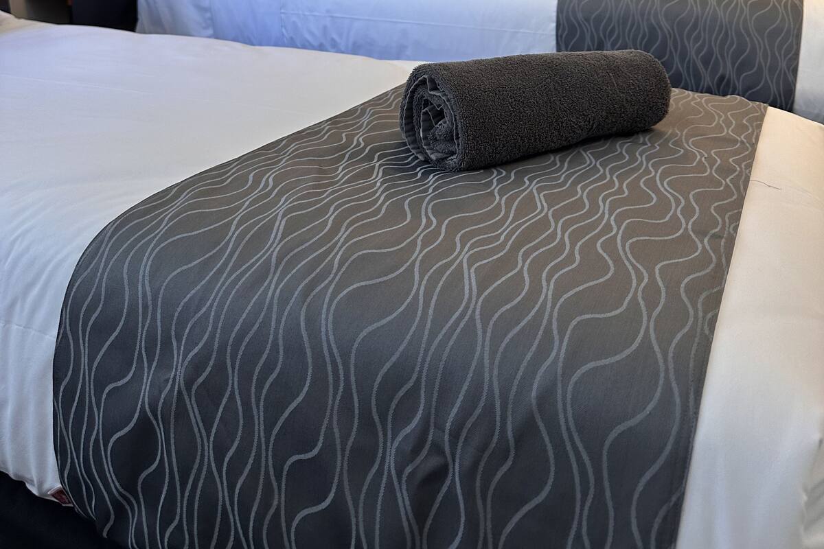 Quarto de hotel com duas camas, cada uma com colcha cinza estampada e uma toalha enrolada. Uma televisão de tela plana está fixada na parede acima das camas, e uma pequena geladeira é visível ao fundo.