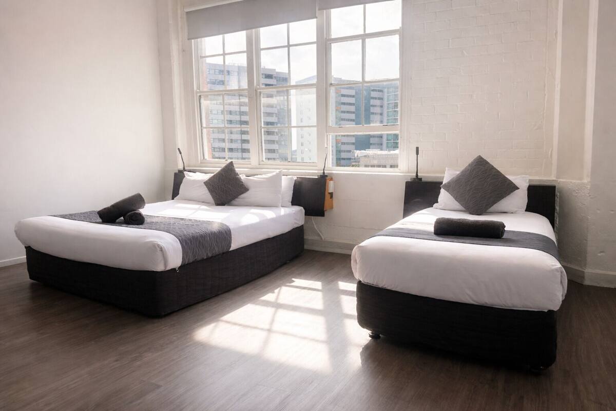 Quarto moderno com duas camas, uma janela grande e parede de tijolos.