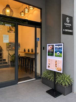 Entrada de hotel com placa indicando 'Gamma Hotel', balcão de recepção, cardápio e plantas em vasos.