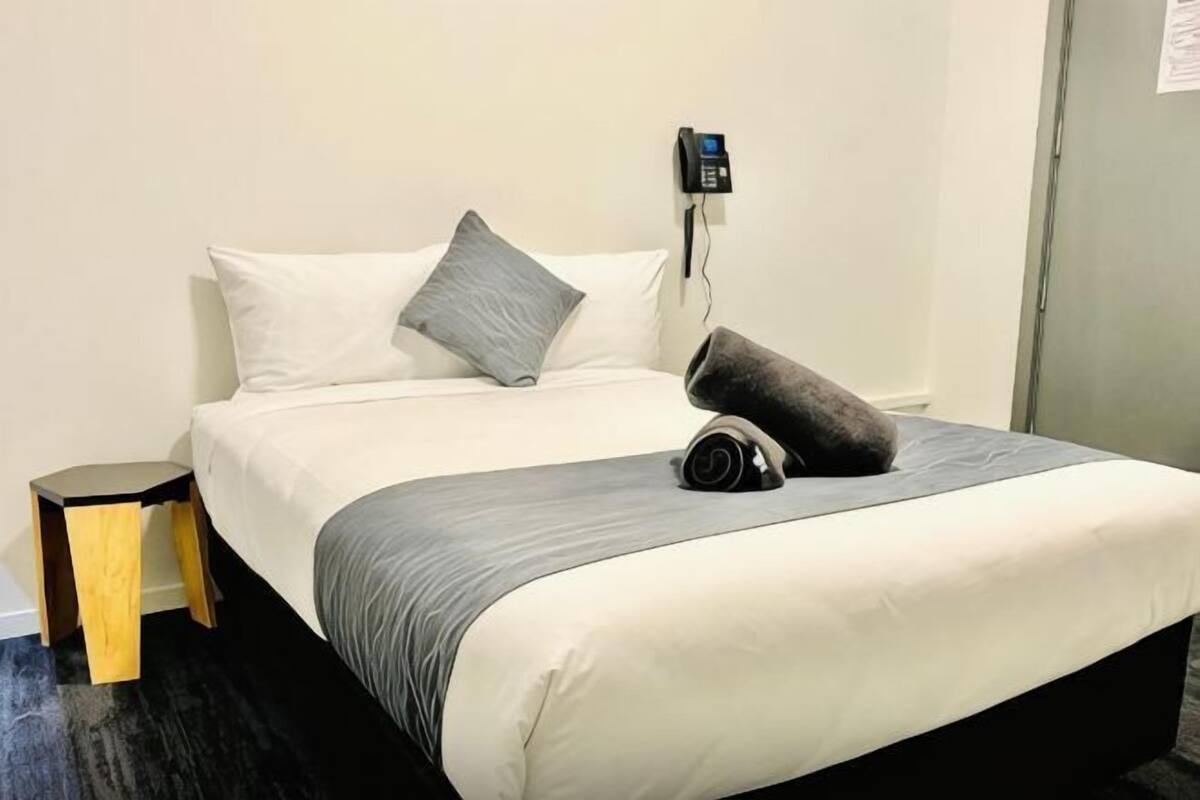 Uma cama bem arrumada com colcha branca e cinza, uma mesinha de cabeceira e um telefone fixado na parede.