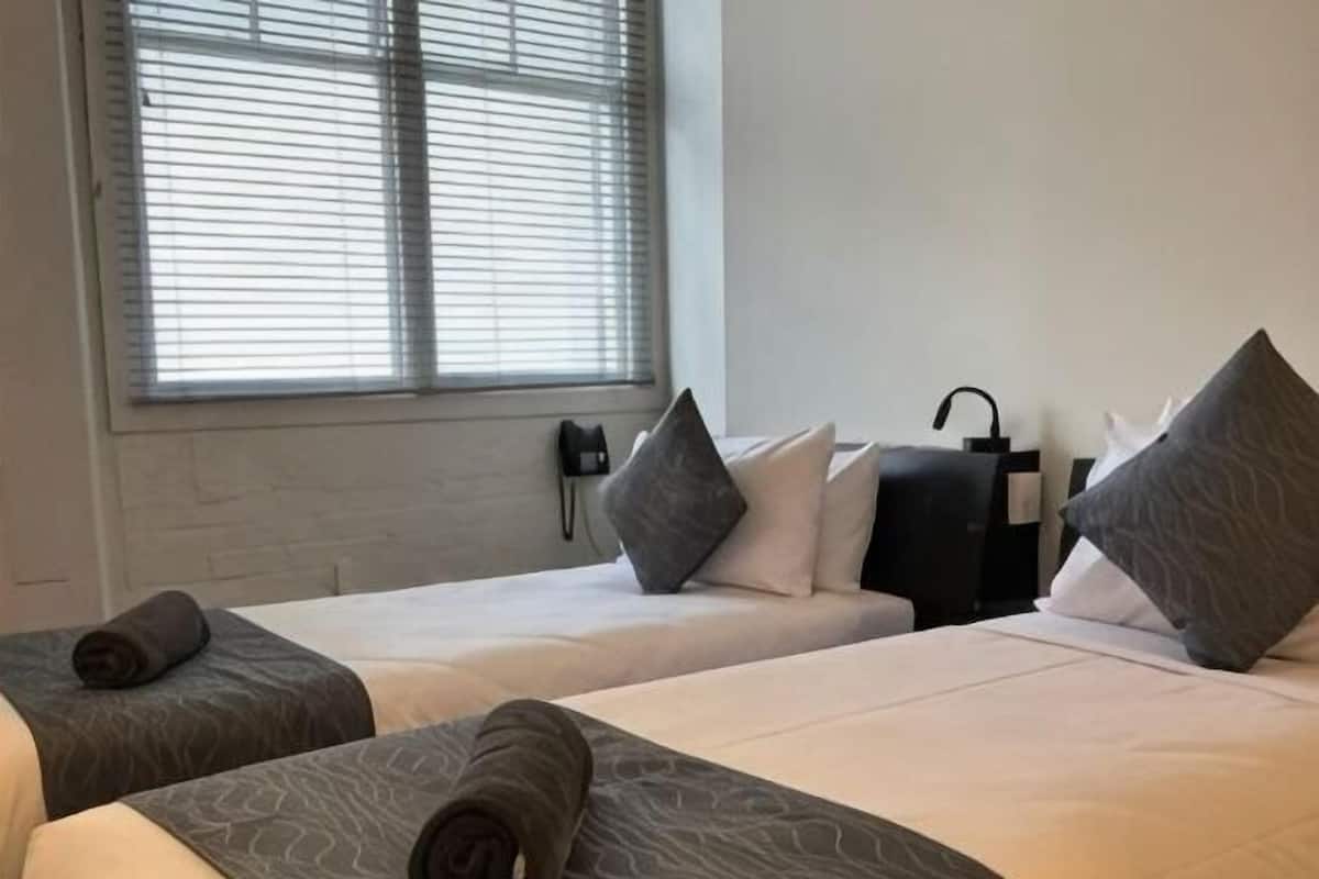 Quarto de hotel com duas camas, janela com persianas, telefone na parede e mala preta no chão.