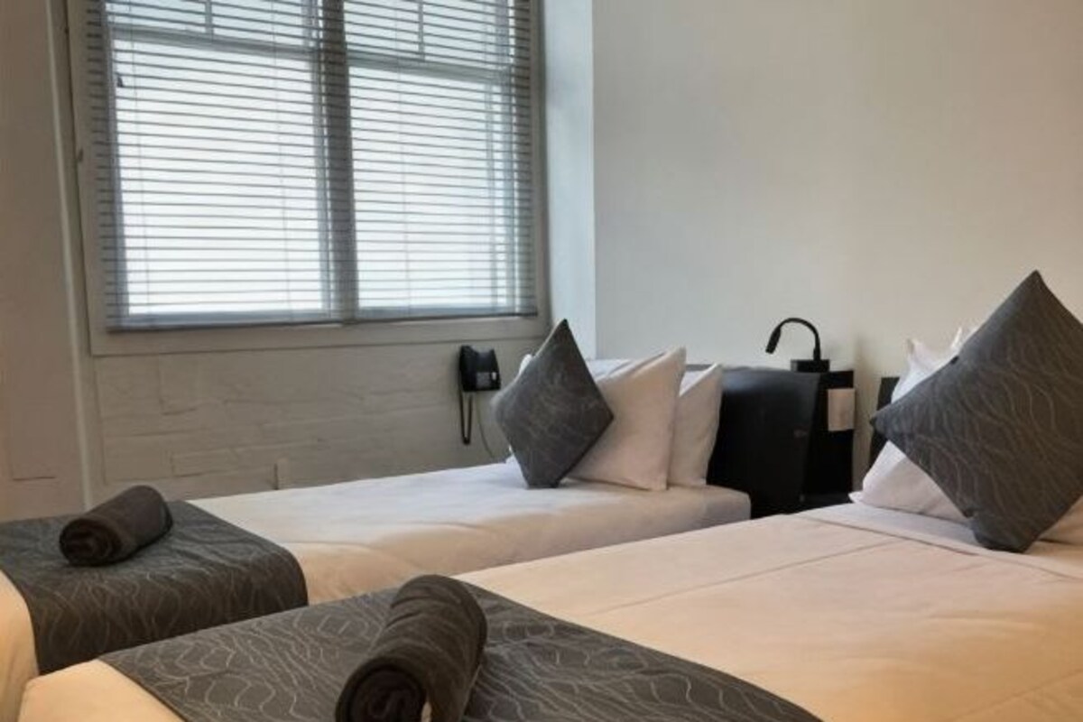 A hotel room with two beds, a window with blinds, a phone on the wall, and a black suitcase on the floor.