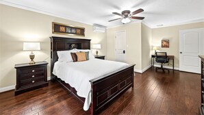 A bedroom with a large bed, two nightstands, a desk with a chair, a ceiling fan, and a door.