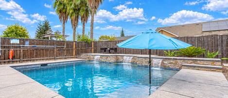 Una piscina rectangular con cascada, rodeada por una valla de madera y una sombrilla azul.
