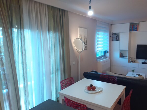 Uma sala de estar moderna com um sofá branco, uma mesa de jantar com uma travessa de frutas, uma cadeira vermelha e uma TV fixada na parede.