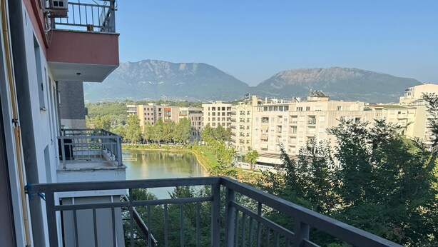 A balcony with two red chairs, a view of a lake, and buildings in the background. - Lake View Apartment (Qarku i Tiranës)