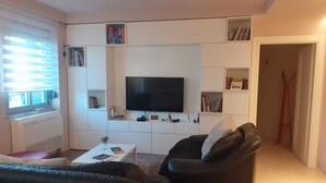 A modern living room with a flat-screen TV, built-in shelves, a white entertainment unit, and a black leather sofa. - Lake View Apartment (Qarku i Tiranës)