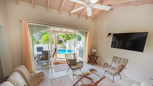 Amplia sala de estar con un gran ventanal corredero que da a una zona de piscina, ventilador de techo y televisor de pantalla plana montado en la pared.