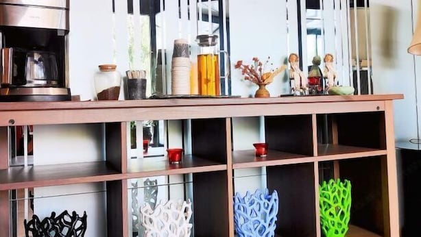 Un coin cuisine équipé d’une machine à café, d’étagères ornées d’objets décoratifs et d’une fenêtre avec des stores verticaux.
