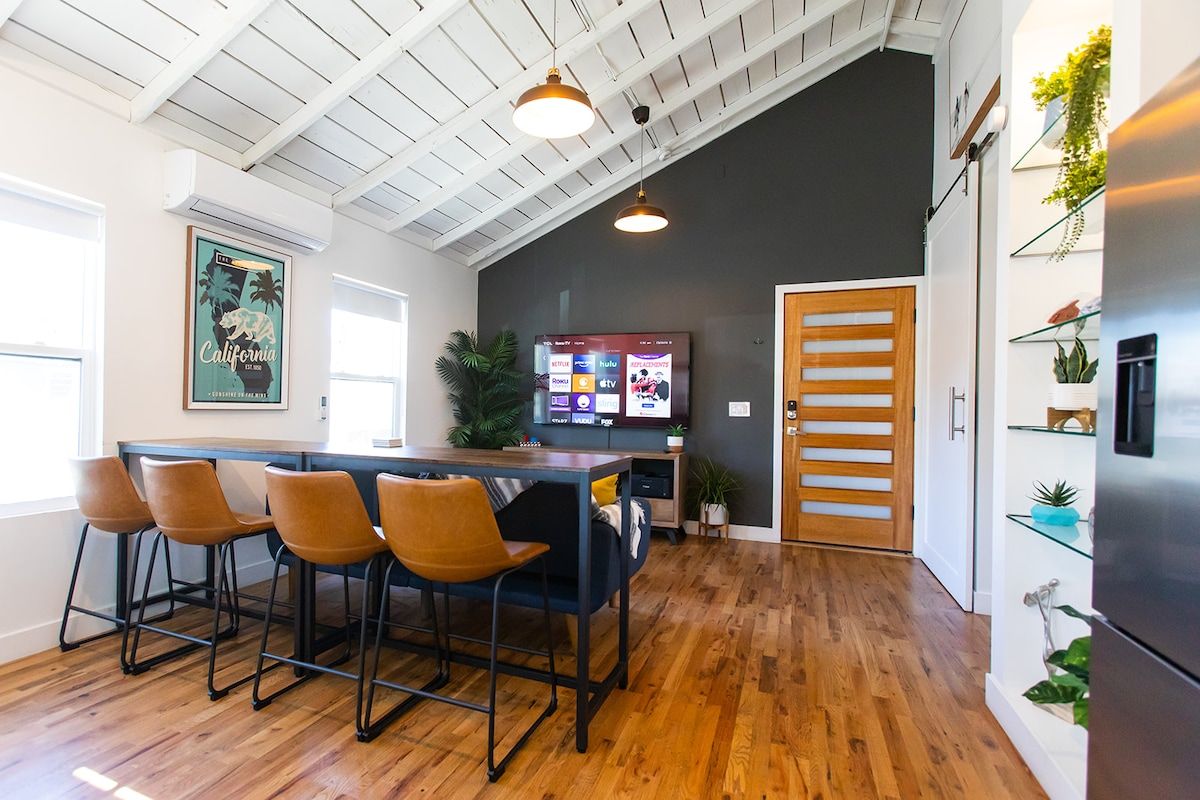 A modern kitchen with a wooden ceiling, a bar area with stools, a flat-screen TV, and a refrigerator.