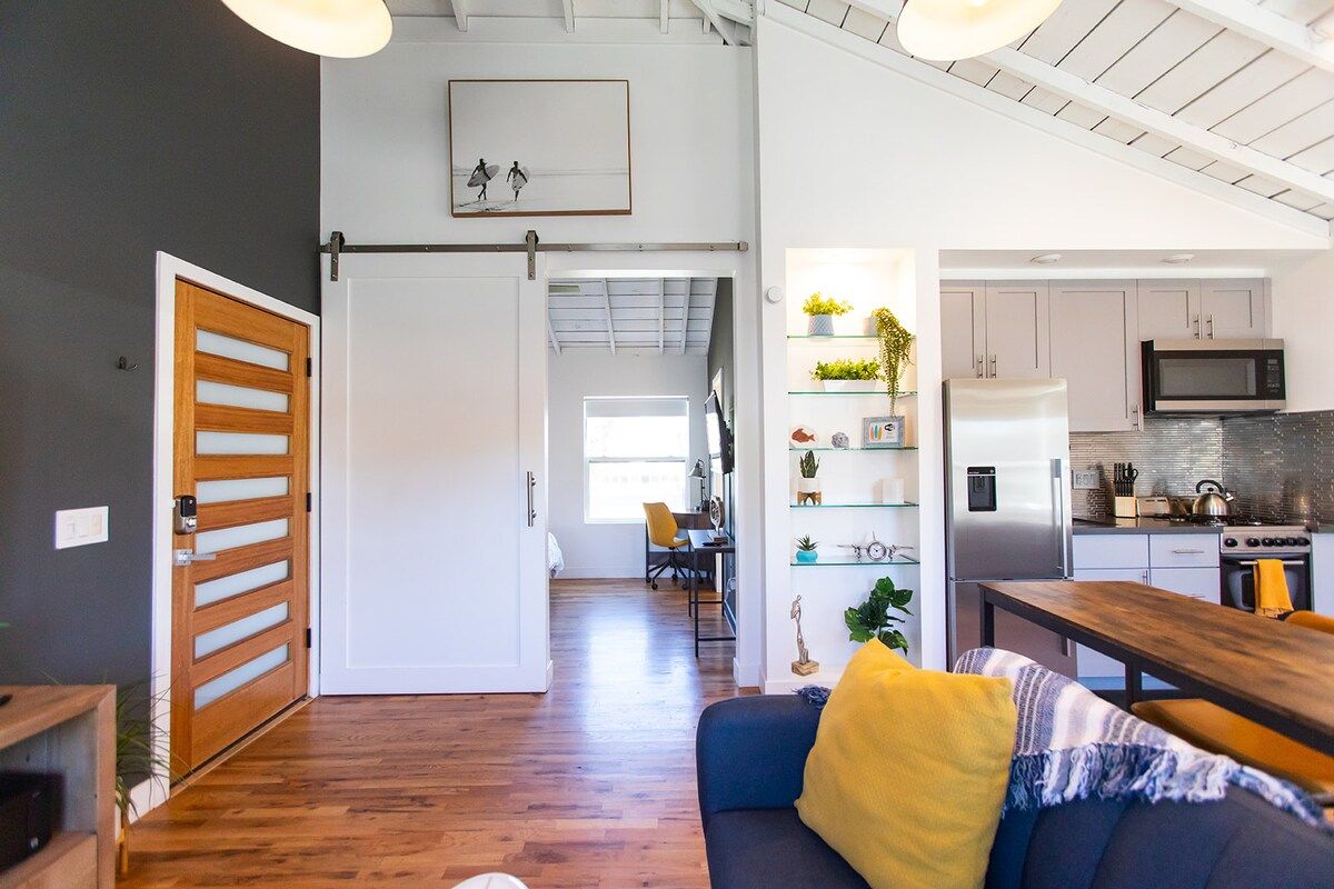 A modern living space with a wooden floor, a sofa with a yellow pillow, a dining table, and a kitchen area with a refrigerator and microwave.