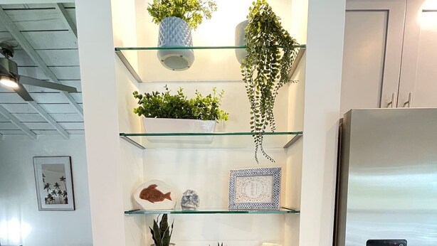 A kitchen with a built-in glass shelf unit holding various decorative items, a refrigerator, and a bed in the background. - Stylish Apt Near Disney and Airport (Santa Ana)