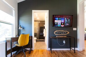 Un salon moderne avec un téléviseur à écran plat fixé au mur, un bureau avec une chaise et un couloir menant à une salle de bain.