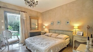 Ein Schlafzimmer mit einem großen Bett, einem Sessel, einem Tisch mit Lampe und einem Fenster mit Vorhängen.