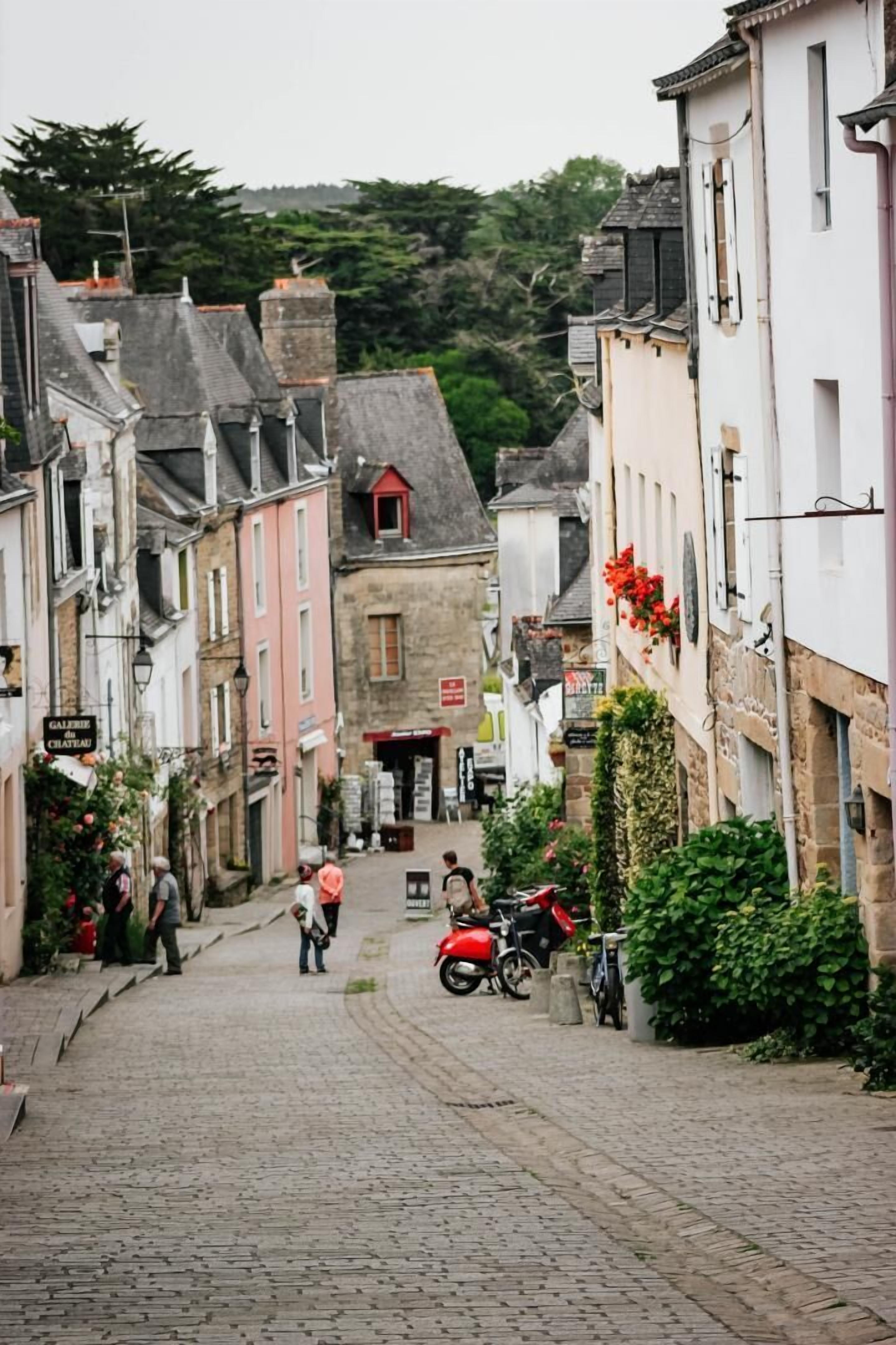 Eine Kopfsteinpflasterstraße in einer historischen Stadt, gesäumt von Gebäuden, mit einem roten Motorrad auf der rechten Seite und einigen Fußgängern.