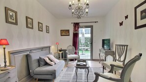 Ein Wohnzimmer mit grauem Sofa, einem kleinen Tisch, einem Fernseher und einem Sessel. Ein Fenster bietet Blick ins Freie.