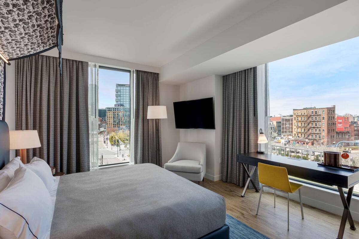A hotel room with a large bed, a desk, a chair, a TV, and a view of the city.