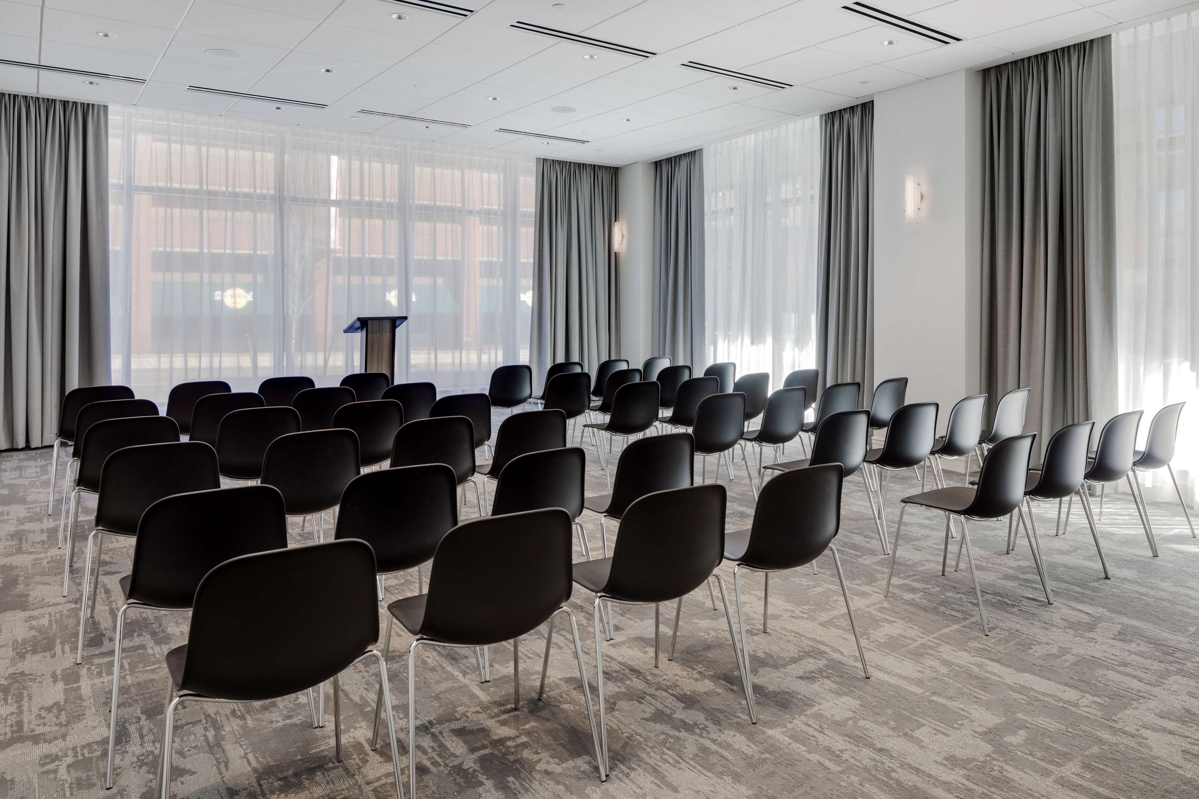 Ett konferensrum med rader av svarta stolar, en talarstol och stora fönster med gardiner.