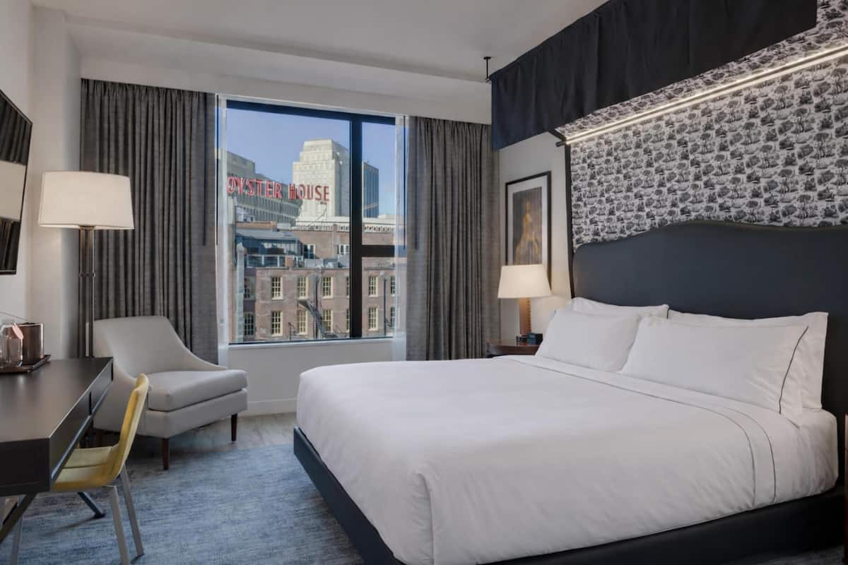 A hotel room with a large bed, a desk with a chair, a window with a view of the city, and a patterned wall.