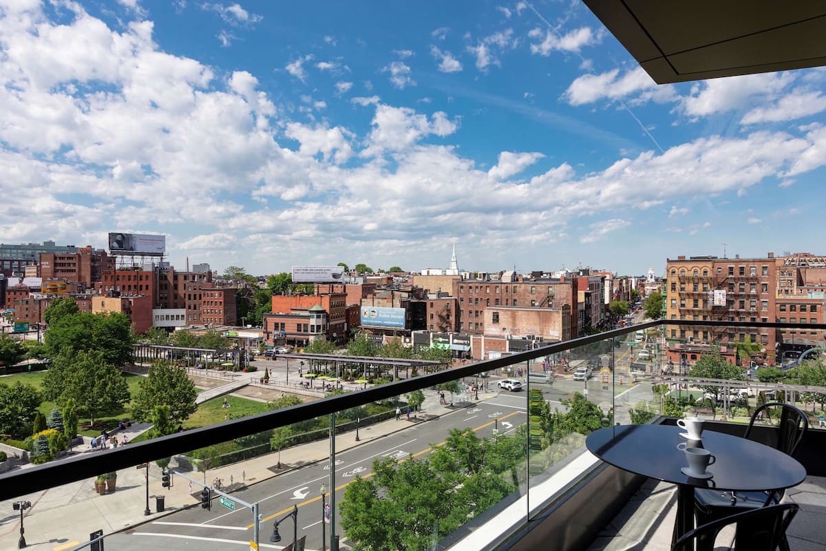 A city view from a balcony with a table and chairs, overlooking a busy street and buildings.