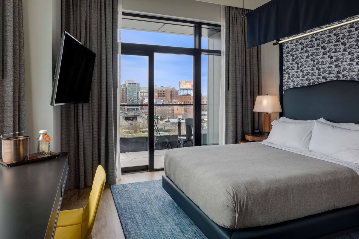 A hotel room with a large bed, a TV mounted on the wall, a desk with a lamp, and a view of the city through the window.