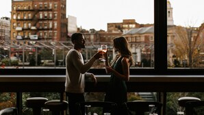 A couple toasting with drinks at a bar with a city view. - Canopy By Hilton Boston Downtown (Boston)