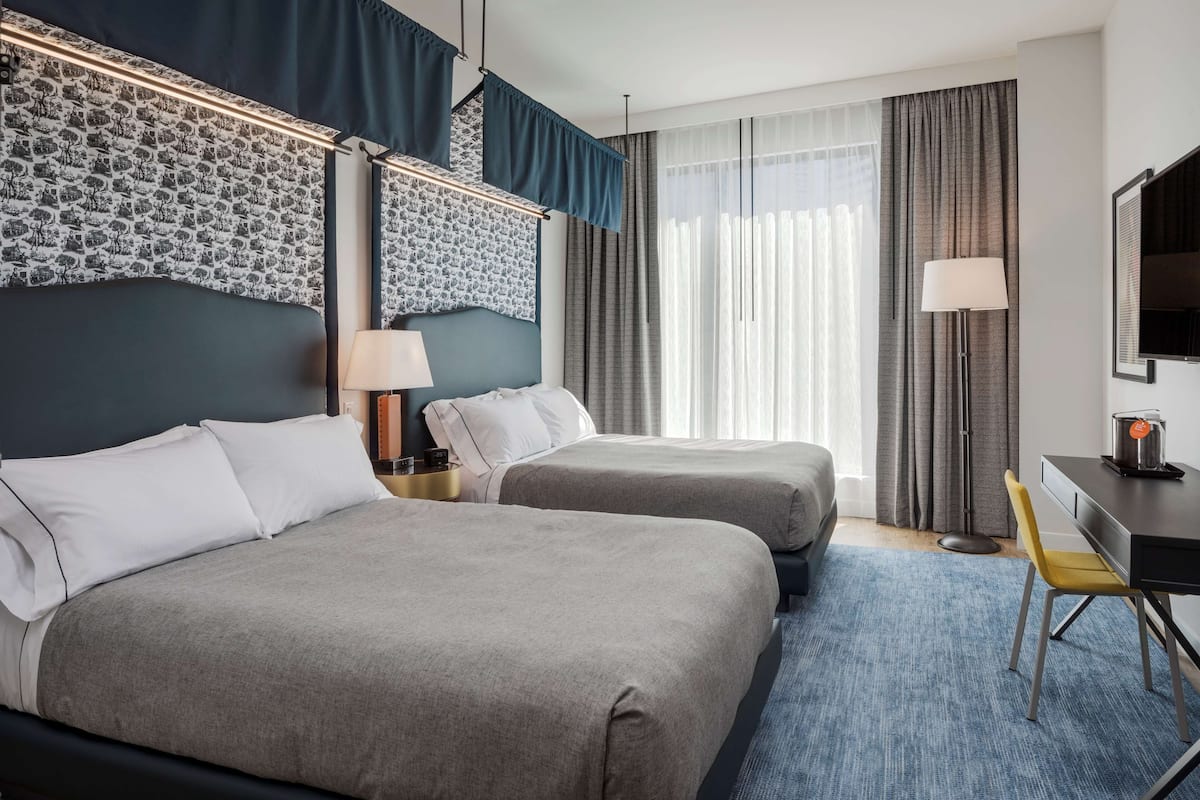 A hotel room with two beds, a desk, a chair, a TV, and a window with curtains.