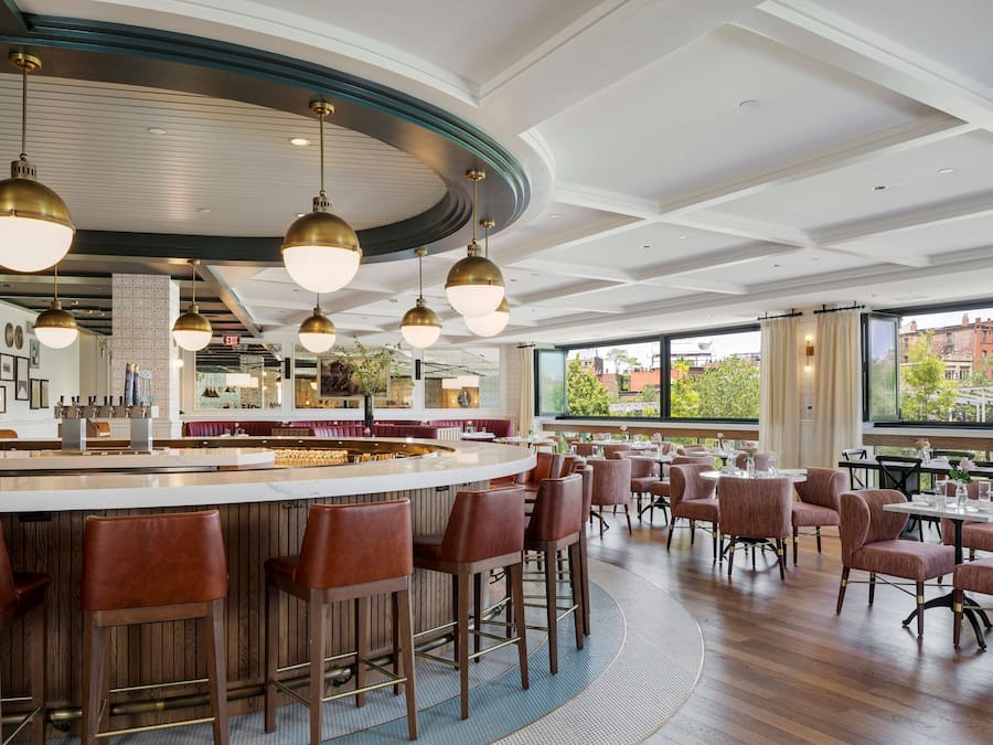 A modern restaurant with a bar area, high ceilings, and large windows offering a view of the cityscape.