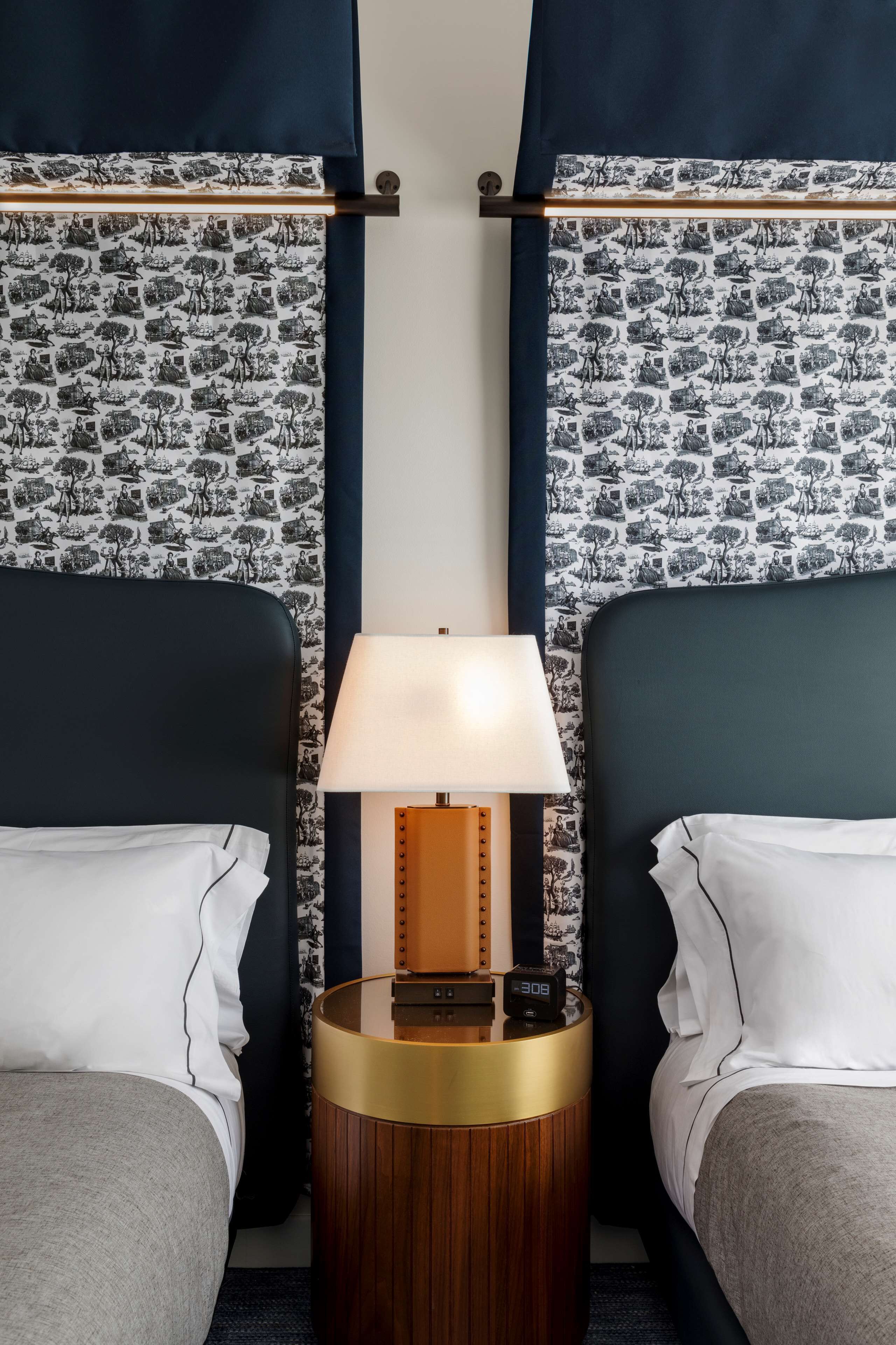 A hotel room with two beds, a nightstand with a lamp, and patterned wallpaper.